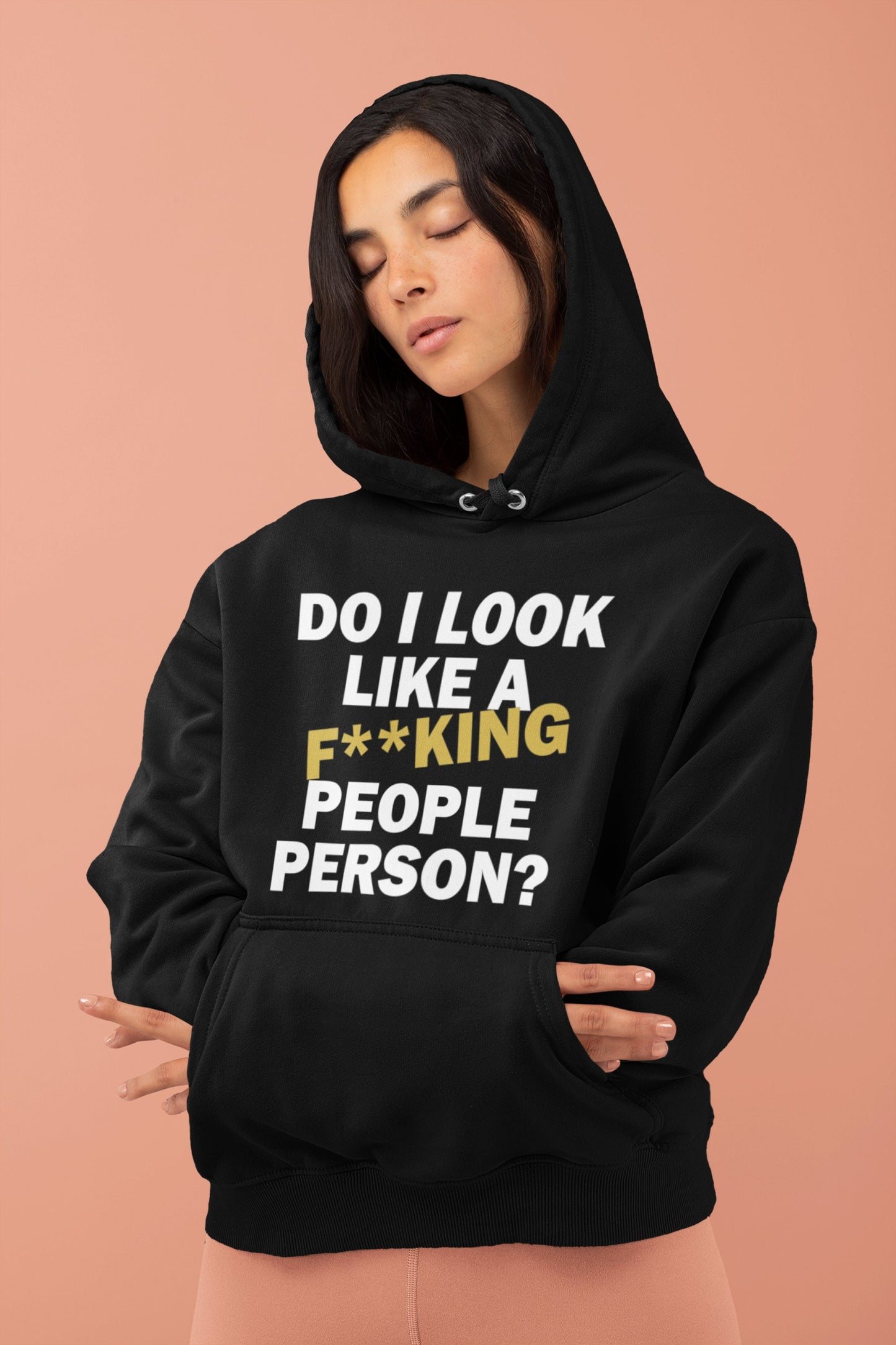 A young woman with long dark hair wearing a black hoodie that reads "Do I Look Like a F**king People Person?" in bold white and gold lettering. The hoodie features a comfortable fit, made from a soft poly-cotton blend, perfect for casual wear or as a humorous gift. The woman stands against a soft pink background with her eyes closed, showcasing the hoodie’s design and relaxed style. The size chart is also displayed, providing detailed measurements for various sizes from S to 4XL.